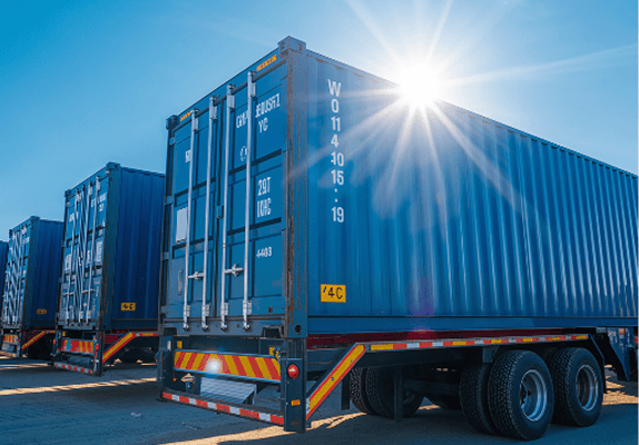 Blue shipping containers on a flatbed trailer, backlit by the sun